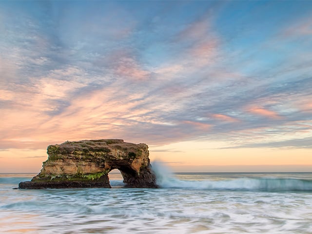 Natural Bridges State beach of waves crashing and a blue pink sky in the background.