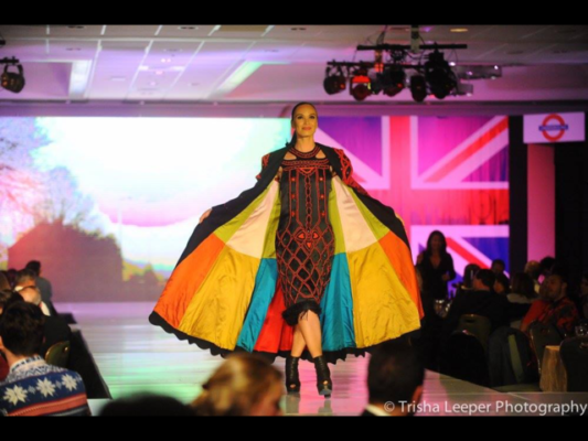 Woman walking down a fashion run way with a multi colored cape on. The colors include, yellow. orange, green, white and teal. The dress she is wearing is black and red.