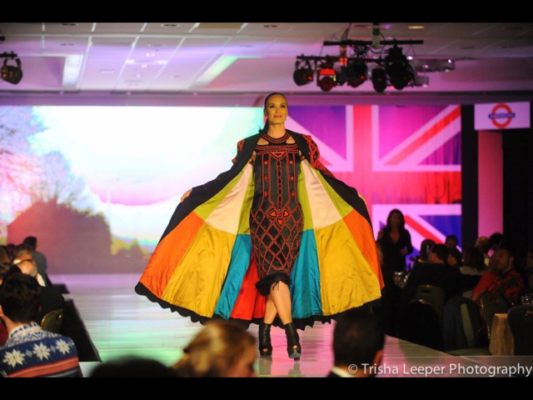 Woman walking down a fashion run way with a multi colored cape on. The colors include, yellow. orange, green, white and teal. The dress she is wearing is black and red. 