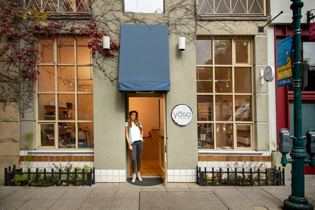 Yoso wellness Spa has a blue awning and the owner is standing underneath it wearing her blonde hair down, a white top, gray pants. They provide facials, massages and acupuncture as well as eco-friendly nail services and more. Their Yoso the Annex provides non-toxic living products.