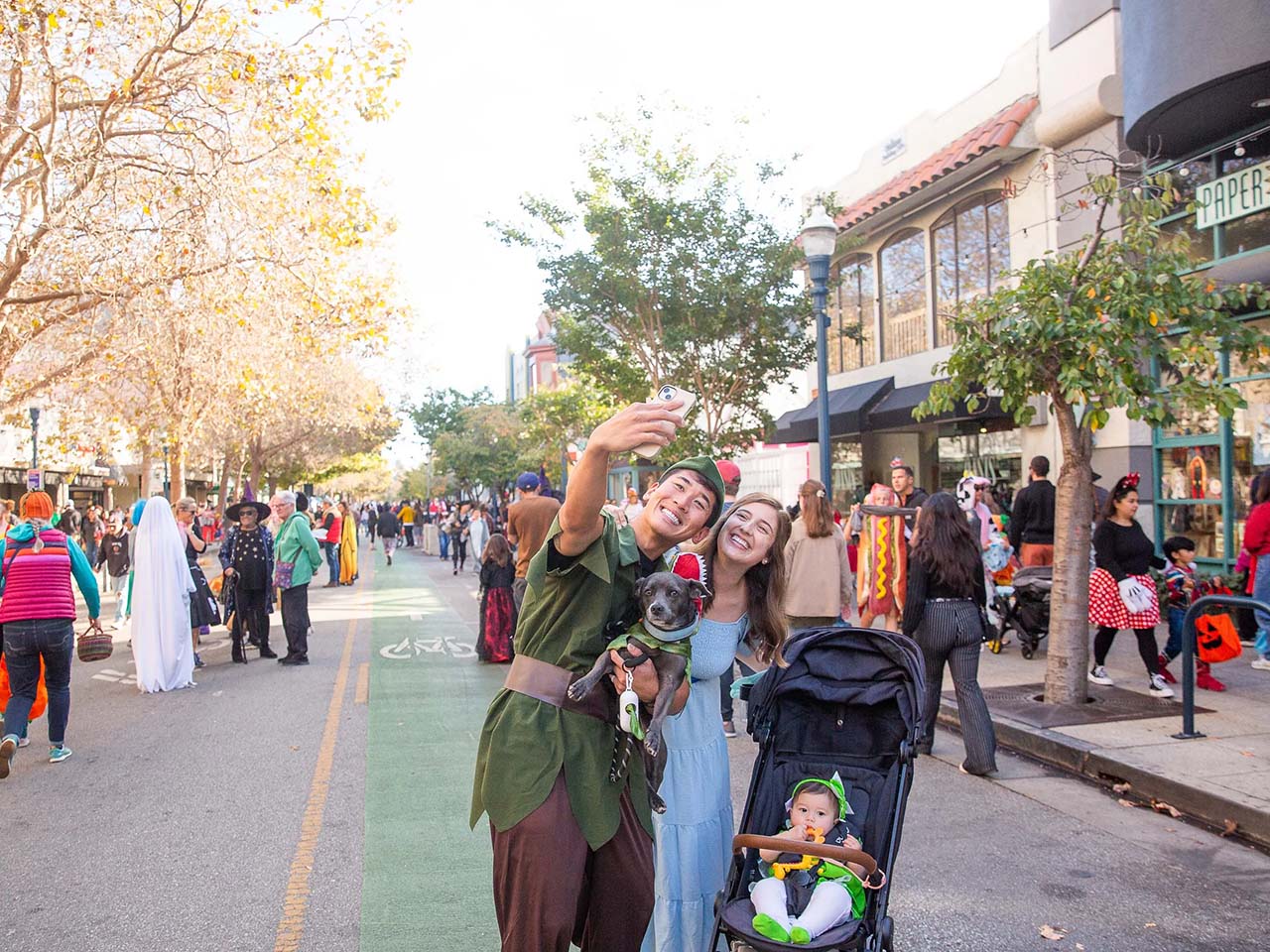 Peter Pan, a Nanna dog and Wendy taking a selfie with their tinker bell in a stroller in the middle of a street that has other costumed families wandering