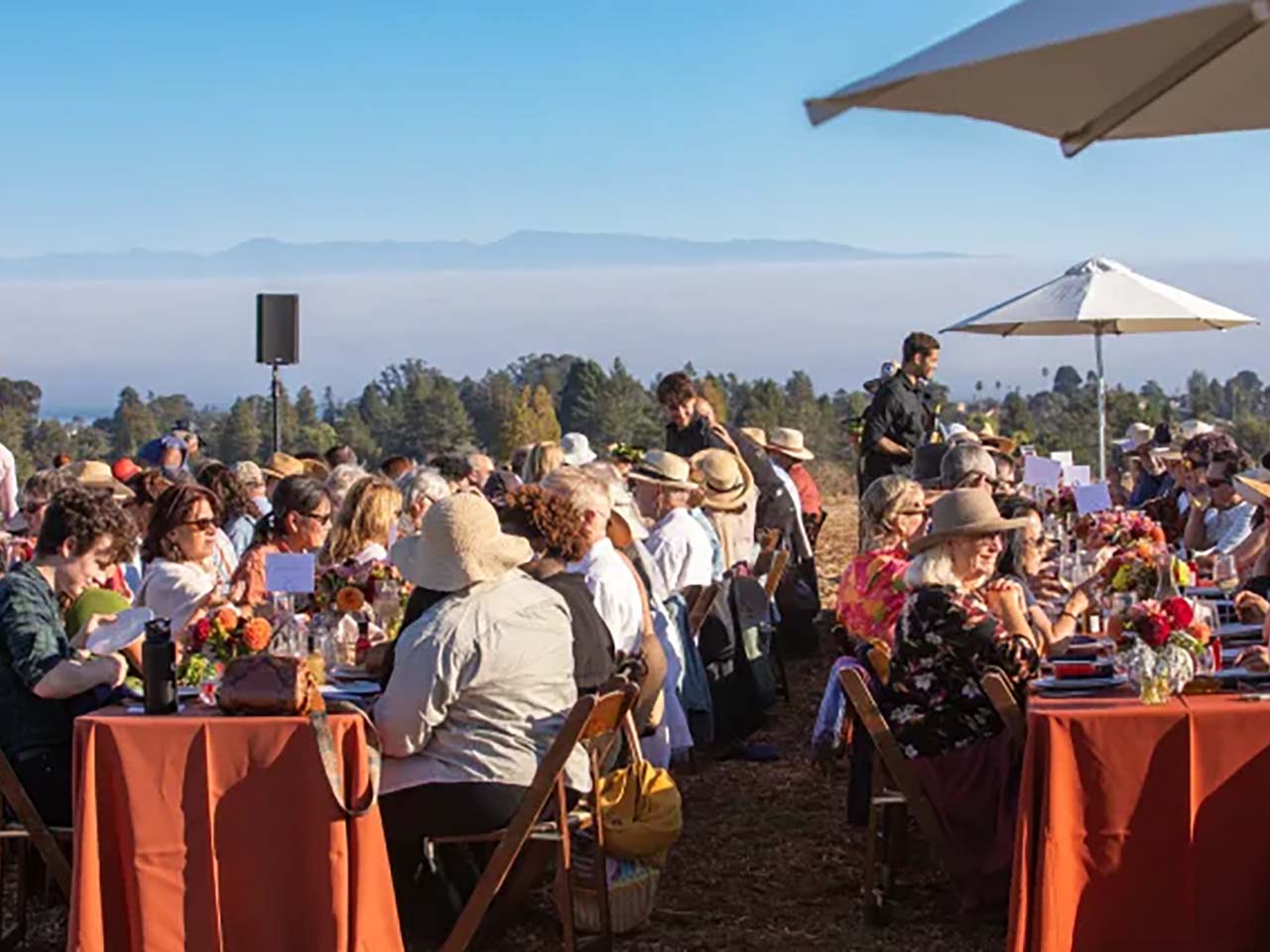 A crowd of people eating at long tables in a farm field with the ocean in the background.