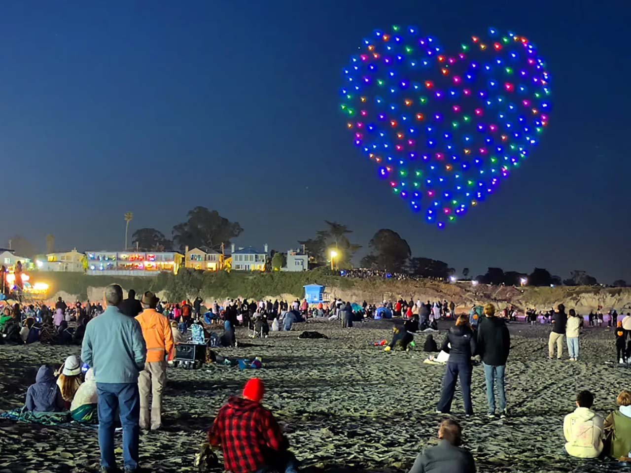 Boardwalk_Winter Opeining Drone Show_2025_1280x960 People bundled up on a sandy beach watching drones form a colorful heart shape in the sky