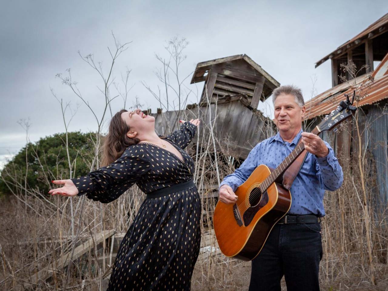 Roaring Camp_Wild and Blue_2025_1280x960 April and Steve Bennett playing guitar and singing by a dilapidated barn with the ocean in the background. Not a Roaring Camp Location.