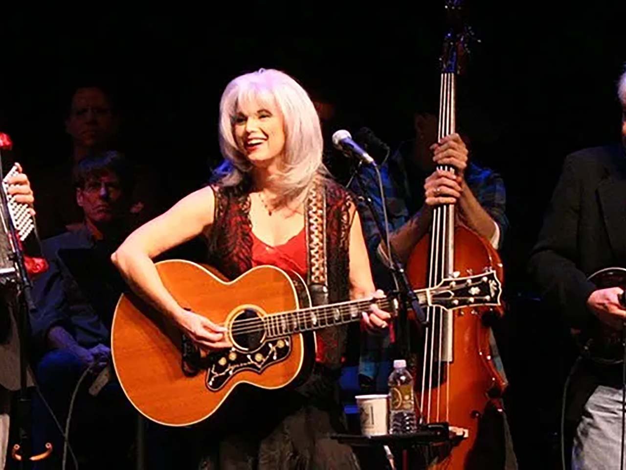 Emmylou on stage with an acoustic guitar, and back up musicians around her.