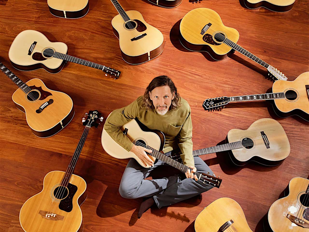Jack west sitting on a floor with acoustic guitars strewn all around him