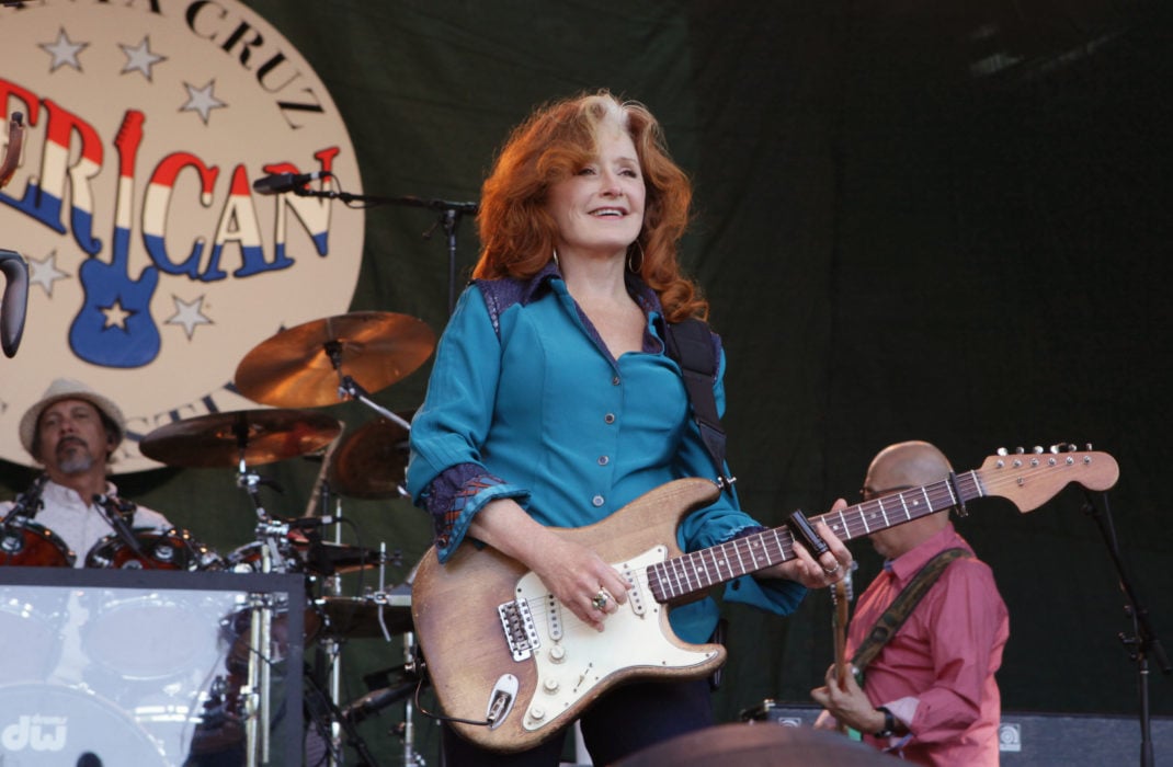 Bonnie Raitt, holding a guitar on stage with a Santa Cruz sign behind her