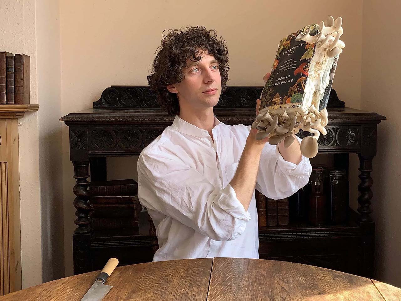Author holding book cover with mushrooms coming out of the edges while a knife is on the table in front of him.