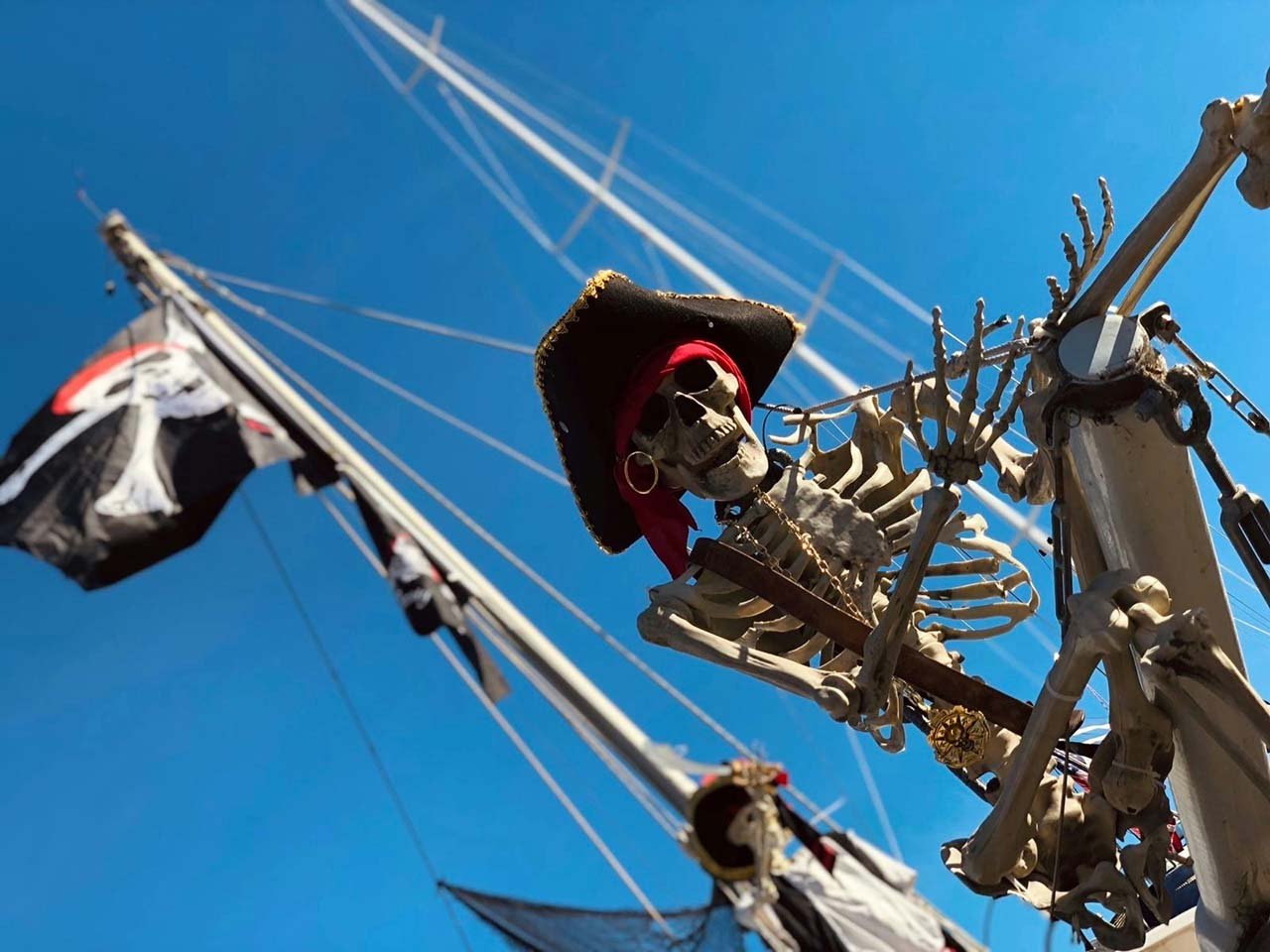 a pirate skeleton looking down from the mast of a boat in the harbor