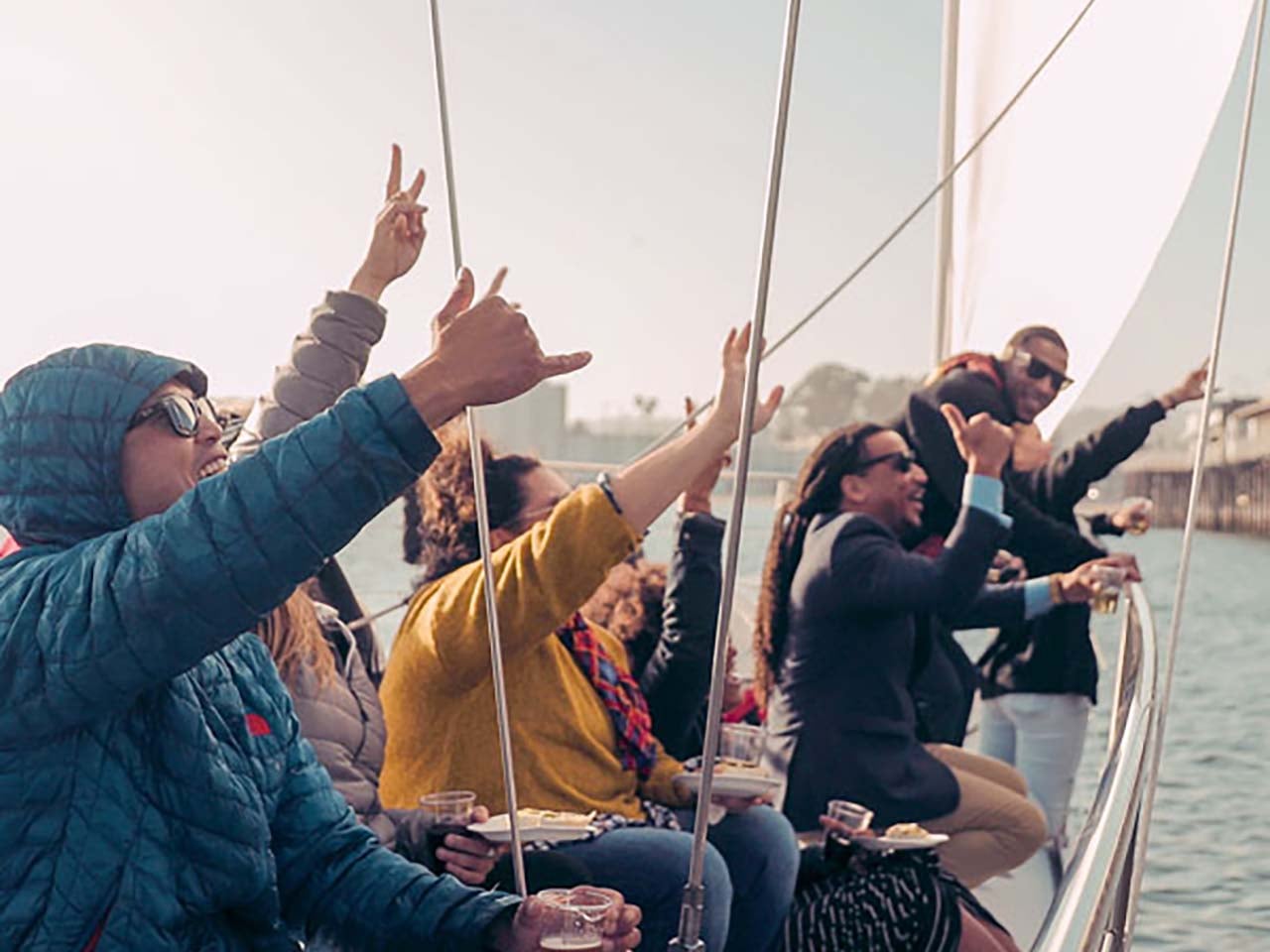 People shaka waving while holding drinks and plates of food on a sailboat