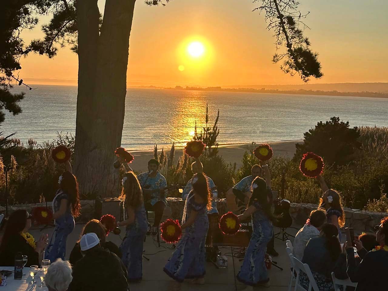 Tahitian dancers performing in front of a sunset overlooking the ocean with an audience around tables.
