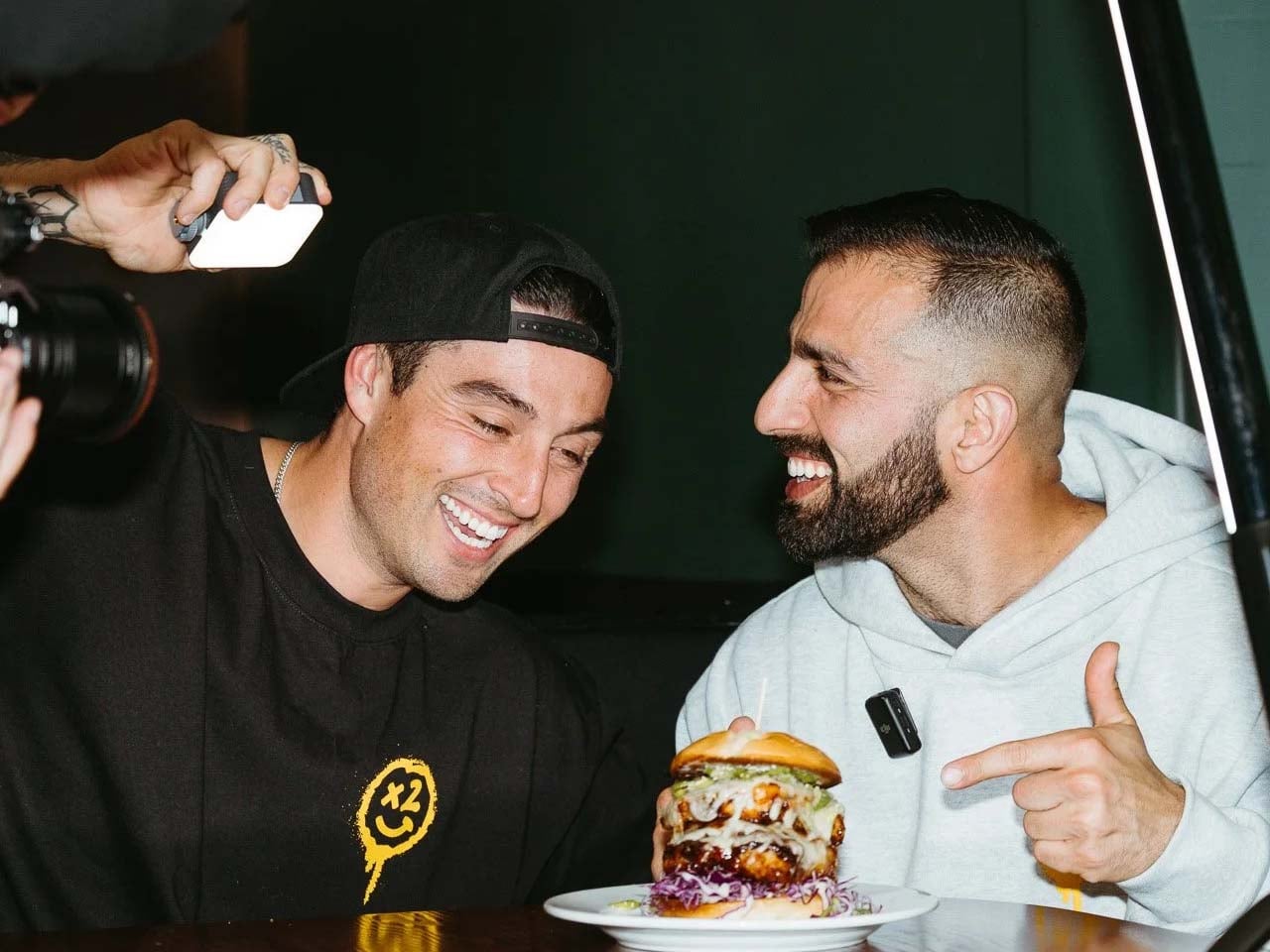 Two food bloggers with camera lights and audio equipment around them smiling and pointing at a gigantic burger on a plate.