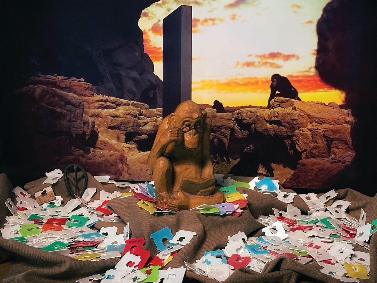 A monolith against a sunset with monkeys around it, and a statue of a thinking monkey with old computer disks around it in the foreground