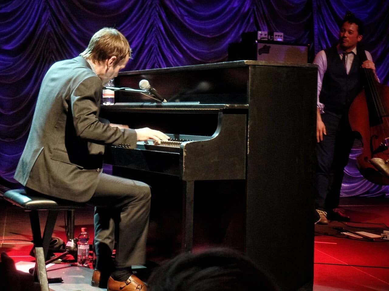 Pianist on an upright piano with a bass player in the background looking on