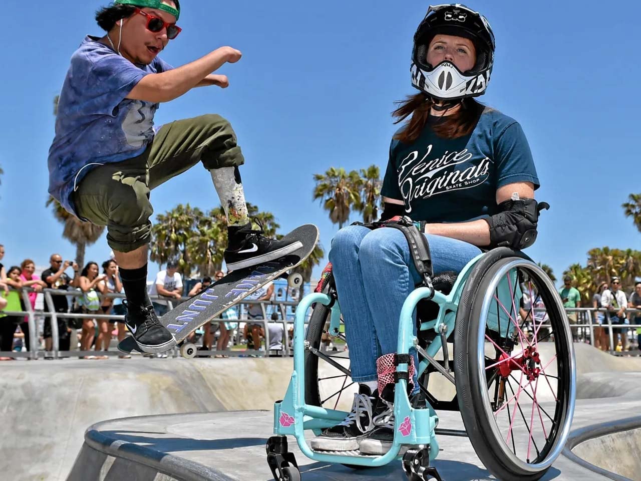 A young man skateboarding with an artificial leg, and a young woman with gear on to navigate the skatepark in her wheelchair