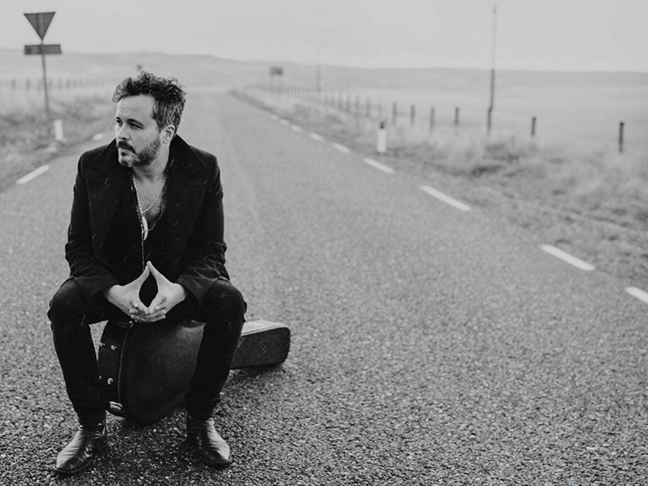 A black and white photo of a man sitting on a guitar case in the middle of an empty road.