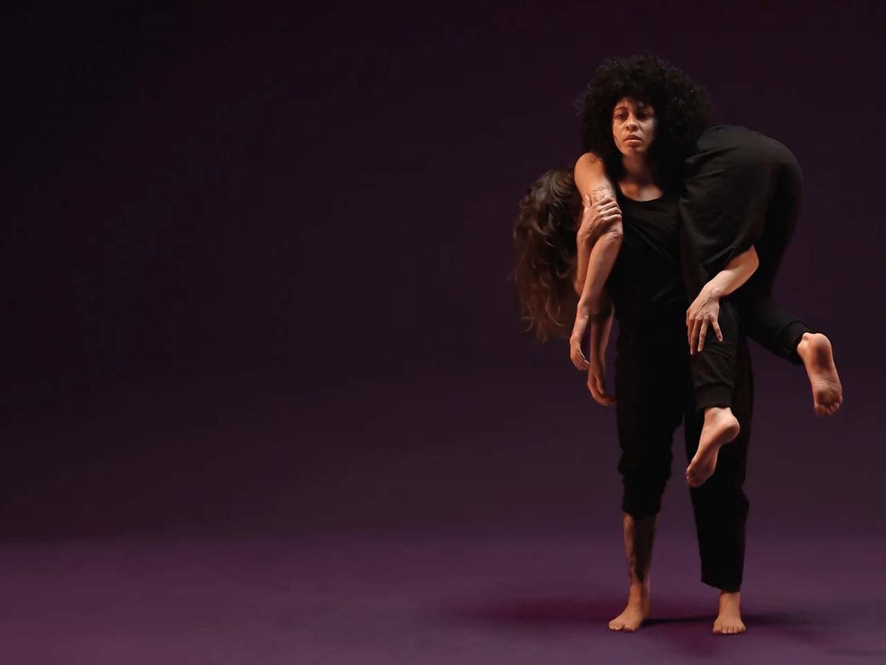 A modern dancer carrying a limp dancer to the far right of a purple background